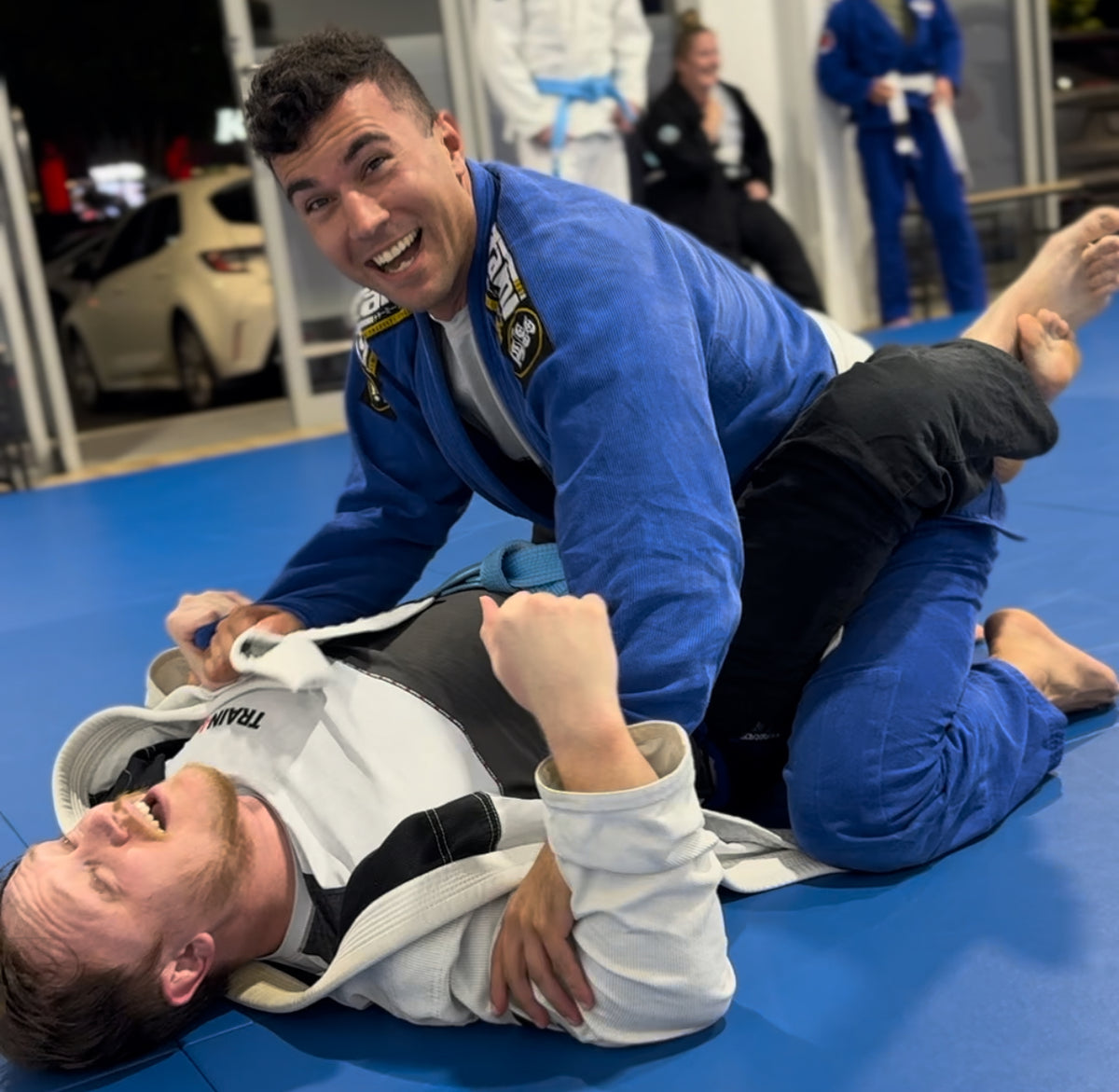 A smiling adult in a blue Gi practising with a training partner at Train4